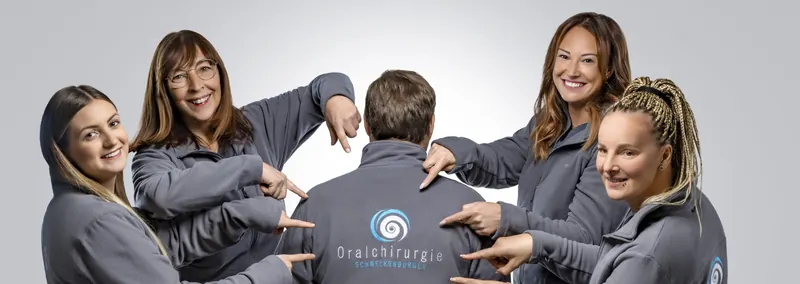 Das Praxisteam zeigt lachend auf das Oralchirurgie-Logo auf dem Rücken eines Kollegen